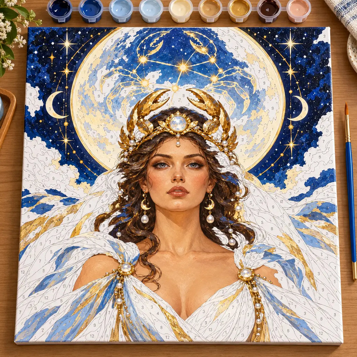 Partially painted Cancer moon goddess paint by numbers canvas with numbered areas, gold crab-claw crown, moonlit sky, and painting supplies.