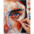 Hands placing diamond drills on an abstract portrait centered on a vivid blue eye.