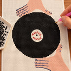 Diamond painting process for Awful Music Vinyl Record Diamond Painting Kit, with drills being placed over a black vinyl record held in two hands, with “Awful Music” at its center.
