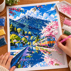 Making the Cherry Blossoms By The Tracks diamond painting, with hands placing drills over pink cherry blossoms lining a mountain railway as a train crosses a blue stream.