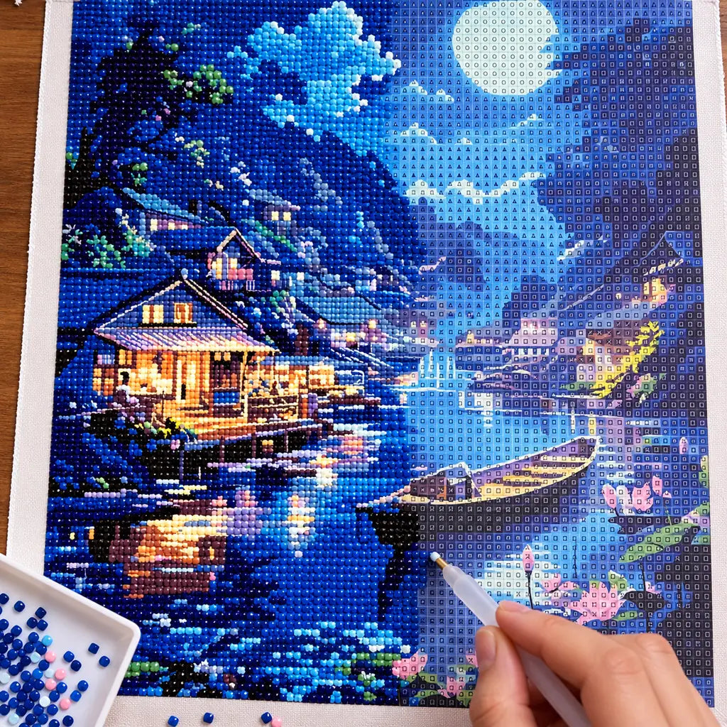 Diamond painting process for Lakeside Moon Glow Diamond Painting Kit, with hands applying drills over a moonlit mountain village and boats reflected on a blue lake.