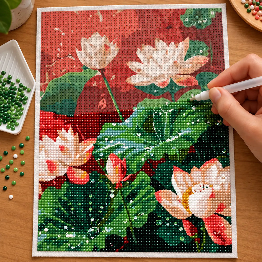 Hands applying drills to a canvas showing white lotus flowers and broad green leaves against a warm red backdrop.