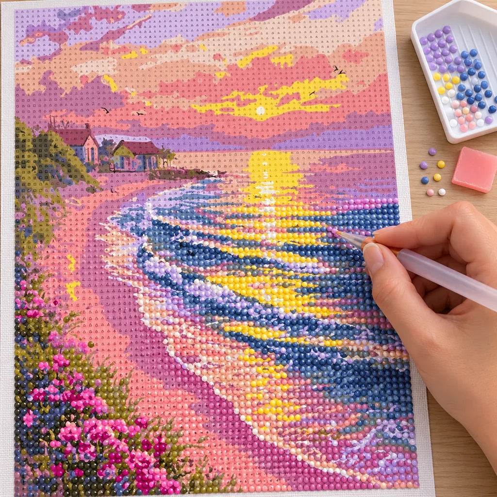 a hand placing diamonds on a pastel sunset over a pink coastal village — process image, diamond painting kit