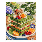 Finished diamond painting showing a bear enjoying fruit beside a matcha layer cake in a leafy picnic setting.