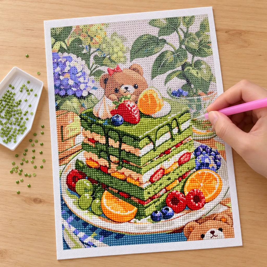 Hands applying drills to a canvas showing a bear enjoying fruit beside a matcha layer cake in a leafy picnic setting.