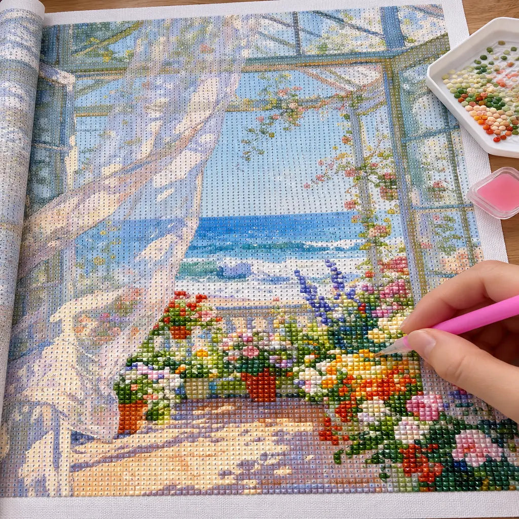 a hand placing diamonds on a flower-filled sunroom by a turquoise sea — process image, diamond painting kit