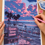 Diamond painting process for Sunset Over Ocean Drive Diamond Painting Kit, with hands applying drills over a palm-lined coastal street beneath pink and lavender sunset clouds.