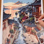 Diamond painting process for Sunset Village by the Stream, showing drills being placed over traditional riverside village homes reflected along a stream beneath sunset clouds.